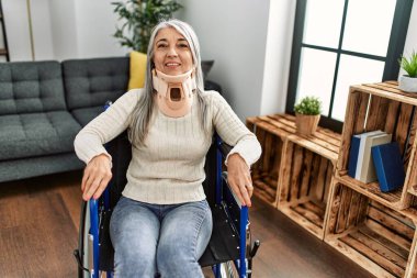 Orta yaşlı, gri saçlı, gülümseyen, kendine güvenen bir kadın, evinde kaza için tekerlekli sandalyede oturuyor.