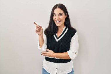 Young hispanic woman standing over isolated background with a big smile on face, pointing with hand and finger to the side looking at the camera. 