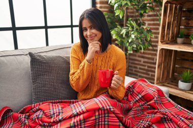 Young hispanic woman sitting on the sofa drinking a coffee at home looking confident at the camera smiling with crossed arms and hand raised on chin. thinking positive. 