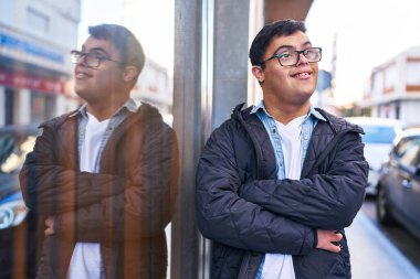 Down sendromlu adam gülümsüyor. Kollarını kavuşturup caddede duruyor.