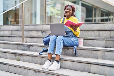 African american woman student using laptop reading book at university