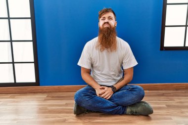 Kızıl saçlı, uzun sakallı bir adam boş bir odada oturmuş komik suratıyla yanaklarını şişiriyor. Havayla şişmiş ağız, çılgın bir ifade.. 