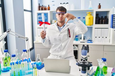 Young hispanic man working at scientist laboratory holding diploma with angry face, negative sign showing dislike with thumbs down, rejection concept 