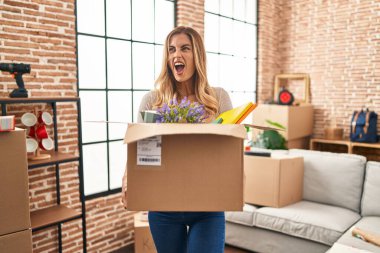 Young blonde woman moving to a new home holding cardboard box angry and mad screaming frustrated and furious, shouting with anger. rage and aggressive concept. 