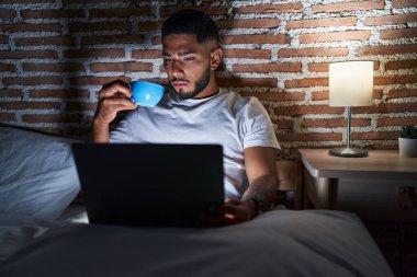 Young latin man drinking coffee using laptop at bedroom