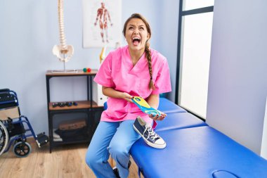 Young hispanic woman holding shoe insole at physiotherapy clinic angry and mad screaming frustrated and furious, shouting with anger looking up. 