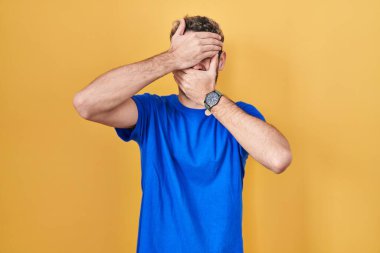 Hispanic man with beard standing over yellow background covering eyes and mouth with hands, surprised and shocked. hiding emotion 