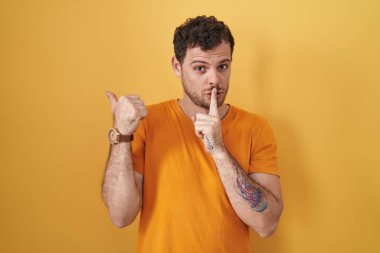 Young hispanic man standing over yellow background asking to be quiet with finger on lips pointing with hand to the side. silence and secret concept. 