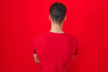 Young hispanic man standing over red background standing backwards looking away with crossed arms 
