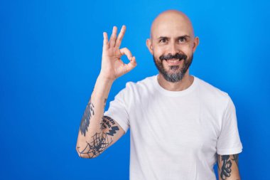 Hispanic man with tattoos standing over blue background smiling positive doing ok sign with hand and fingers. successful expression. 