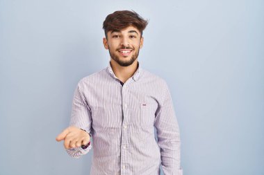 Arab man with beard standing over blue background smiling cheerful offering palm hand giving assistance and acceptance. 