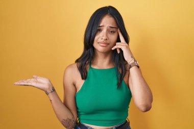 Brunette woman standing over yellow background confused and annoyed with open palm showing copy space and pointing finger to forehead. think about it. 