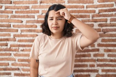 Young hispanic woman standing over bricks wall pointing unhappy to pimple on forehead, ugly infection of blackhead. acne and skin problem 
