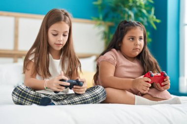 Two kids playing video game sitting on bed at bedroom