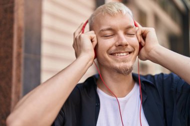 Genç, beyaz bir adam gülerek sokakta müzik dinliyor.