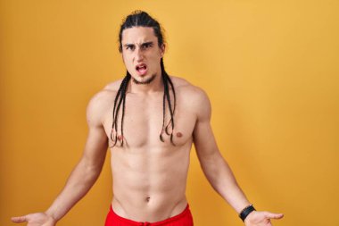 Hispanic man with long hair standing shirtless over yellow background in shock face, looking skeptical and sarcastic, surprised with open mouth 