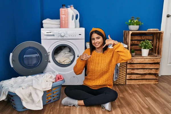Young hispanic woman doing laundry smiling doing talking on the telephone gesture and pointing to you. call me.