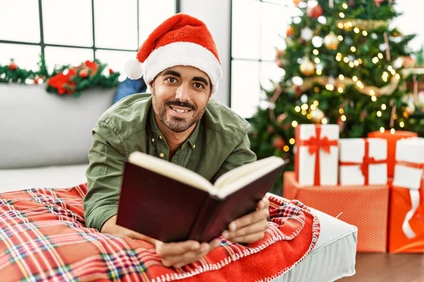 Young Hispanic Man Reading Book Lying Sofa Christmas Tree Home — Φωτογραφία Αρχείου