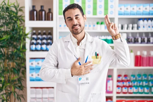 Handsome hispanic man working at pharmacy drugstore smiling swearing with hand on chest and fingers up, making a loyalty promise oath