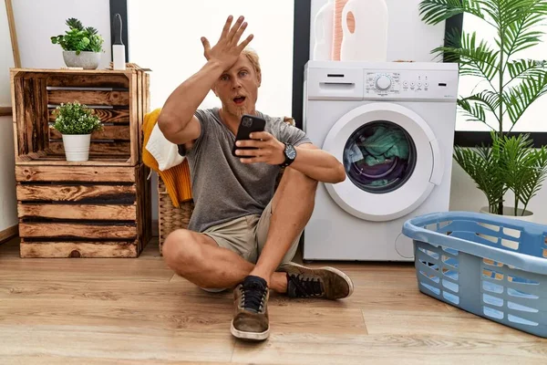 Young Blond Man Doing Laundry Using Smartphone Surprised Hand Head — Stok fotoğraf