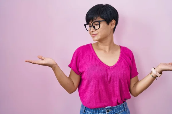 Young asian woman with short hair standing over pink background smiling showing both hands open palms, presenting and advertising comparison and balance