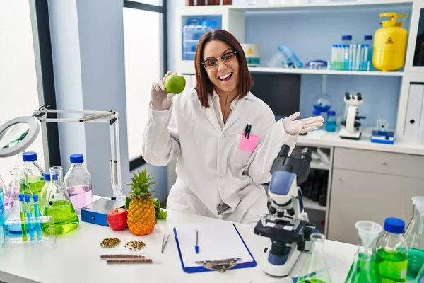 Young Hispanic Woman Working Scientist Laboratory Holding Fruit Celebrating Achievement — Stockfoto