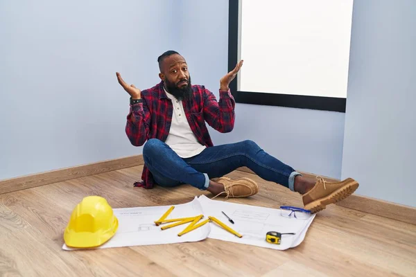 アフリカ系アメリカ人の男性が新しい家を見て床に座っていると 腕や手で発生した設計図を目に見えず 混乱した表情 疑わしい概念 — ストック写真