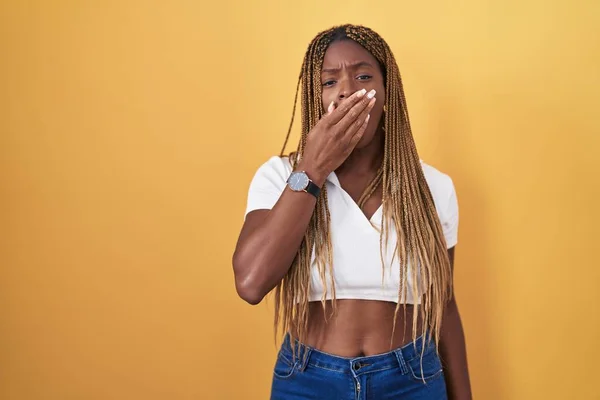 African american woman with braided hair standing over yellow background bored yawning tired covering mouth with hand. restless and sleepiness.