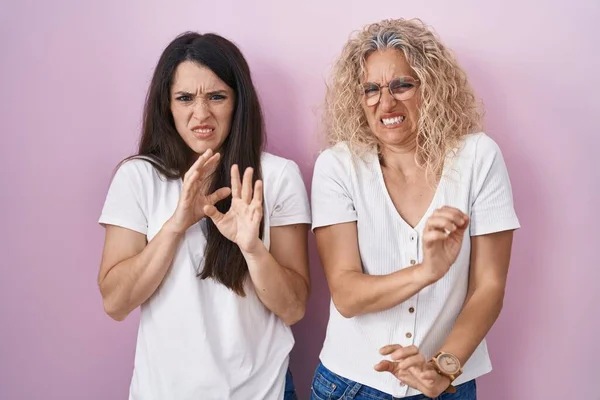 Mother and daughter standing together over pink background disgusted expression, displeased and fearful doing disgust face because aversion reaction.