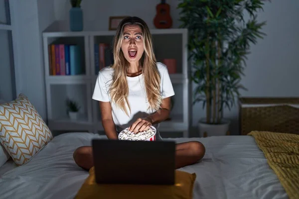 Young blonde woman sitting on the bed at home watching a movie from laptop angry and mad screaming frustrated and furious, shouting with anger looking up.
