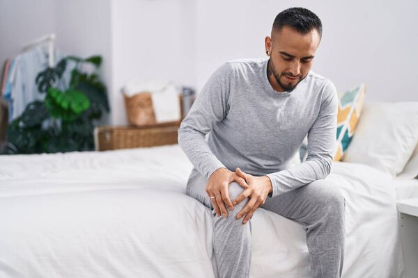 Young hispanic man suffering for knee injury sitting on bed at bedroom