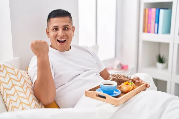 Young Hispanic Man Eating Breakfast Bed Screaming Proud Celebrating Victory — Stockfoto