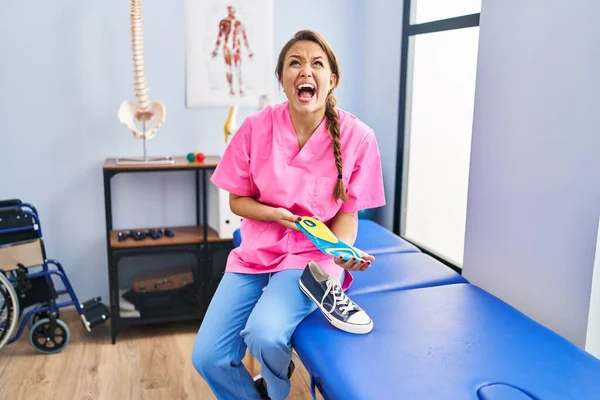 Young hispanic woman holding shoe insole at physiotherapy clinic angry and mad screaming frustrated and furious, shouting with anger looking up.