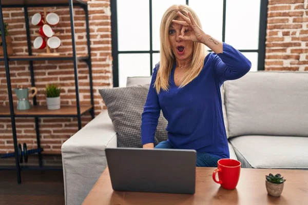 Middle age blonde woman using computer laptop at home doing ok gesture shocked with surprised face, eye looking through fingers. unbelieving expression.