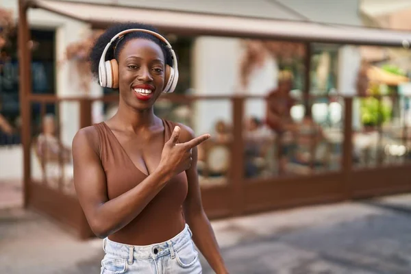 African american woman listening to music pointing with finger to the side at street