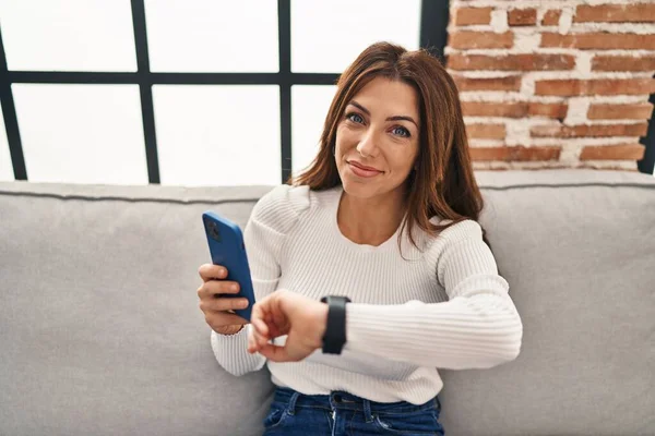 Young hispanic woman using smartphone looking watch at home