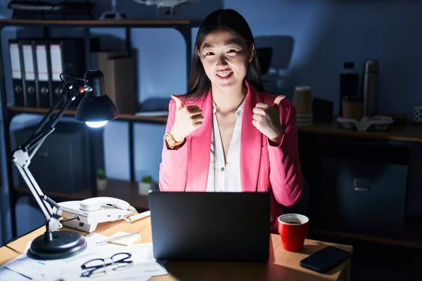 Chinese young woman working at the office at night success sign doing positive gesture with hand, thumbs up smiling and happy. cheerful expression and winner gesture.