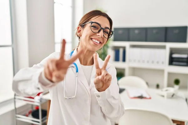 Young brunette doctor woman wearing stethoscope at the clinic smiling looking to the camera showing fingers doing victory sign. number two.