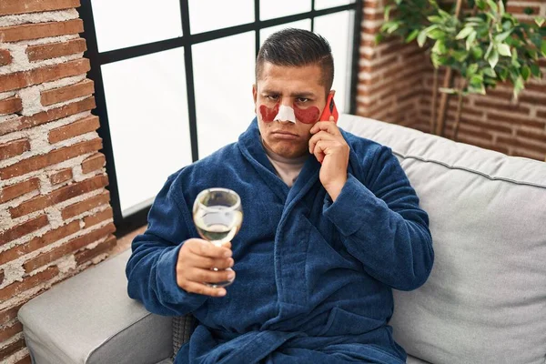 Hispanic young man wearing bathrobe and eye bags patches drinking wine speaking on the phone depressed and worry for distress, crying angry and afraid. sad expression.