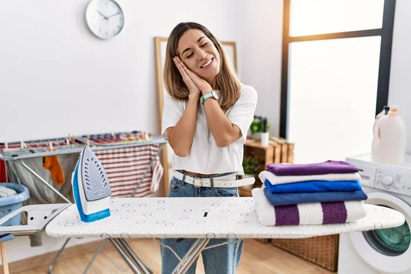 stock image Young hispanic woman ironing clothes at laundry room sleeping tired dreaming and posing with hands together while smiling with closed eyes. 