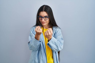 Young hispanic woman standing over blue background ready to fight with fist defense gesture, angry and upset face, afraid of problem 