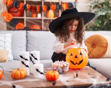 Adorable hispanic girl having halloween party putting sweets on pumpkin basket at home