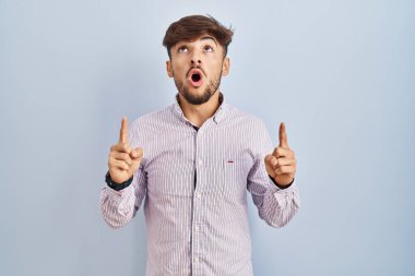 Arab man with beard standing over blue background amazed and surprised looking up and pointing with fingers and raised arms. 