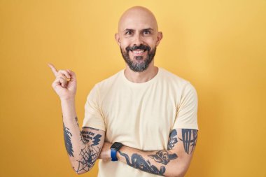 Hispanic man with tattoos standing over yellow background with a big smile on face, pointing with hand finger to the side looking at the camera. 