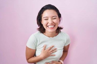 Hispanic young woman standing over pink background smiling and laughing hard out loud because funny crazy joke with hands on body. 