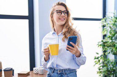 Genç sarışın iş kadını akıllı telefon kullanıyor. Ofiste kahve içiyor.