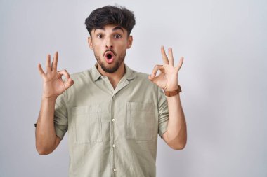 Arab man with beard standing over white background looking surprised and shocked doing ok approval symbol with fingers. crazy expression 