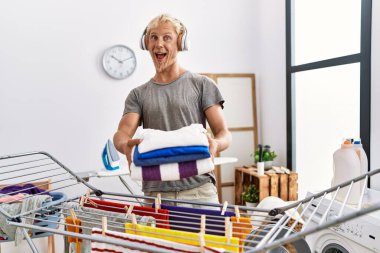 Young blond man listening to music doing laundry celebrating crazy and amazed for success with open eyes screaming excited. 