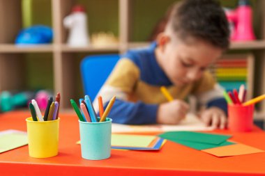 Adorable hispanic boy preschool student sitting on table drawing on paper at kindergarten