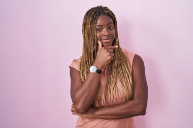 African american woman with braided hair standing over pink background looking confident at the camera smiling with crossed arms and hand raised on chin. thinking positive. 